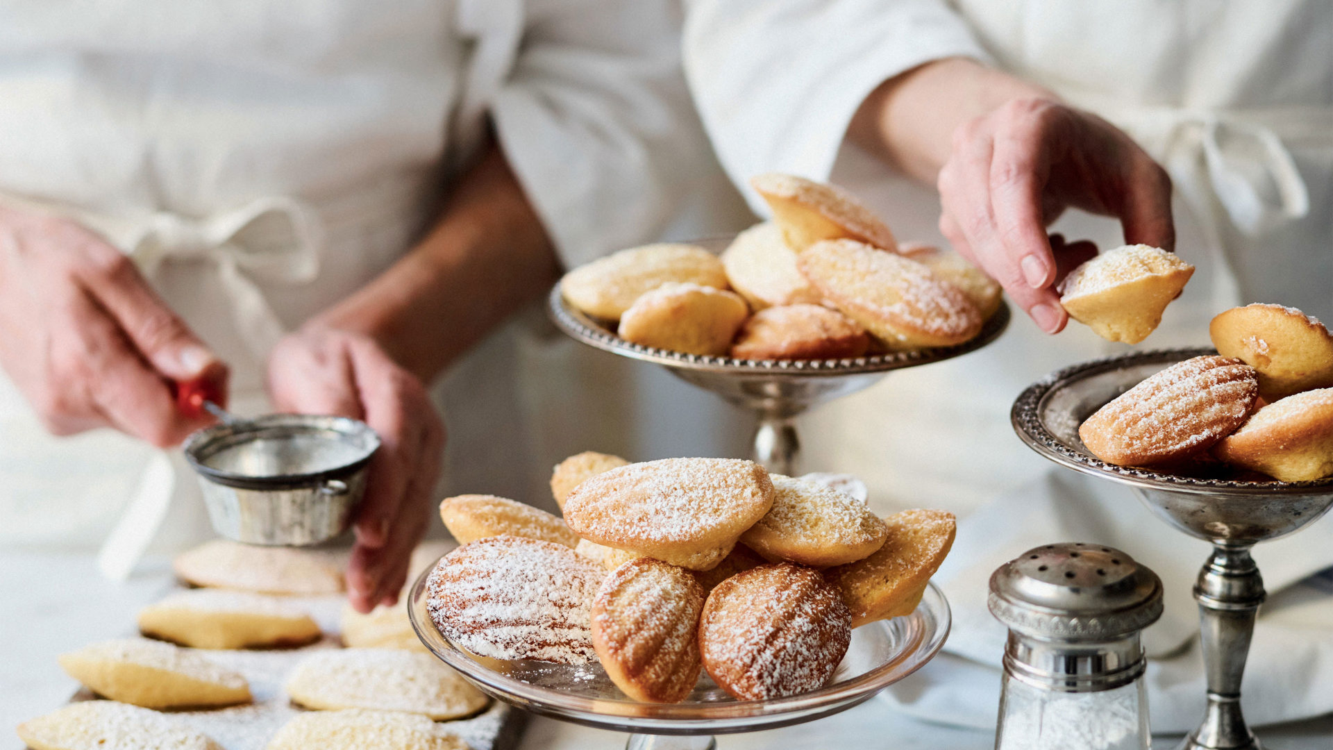 Two chefs prepare and dust dishes of madeleines with sugar