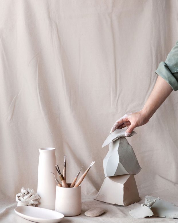 A hand arranges ceramic sculptures alongside artist tools against a pastel pink studio backdrop